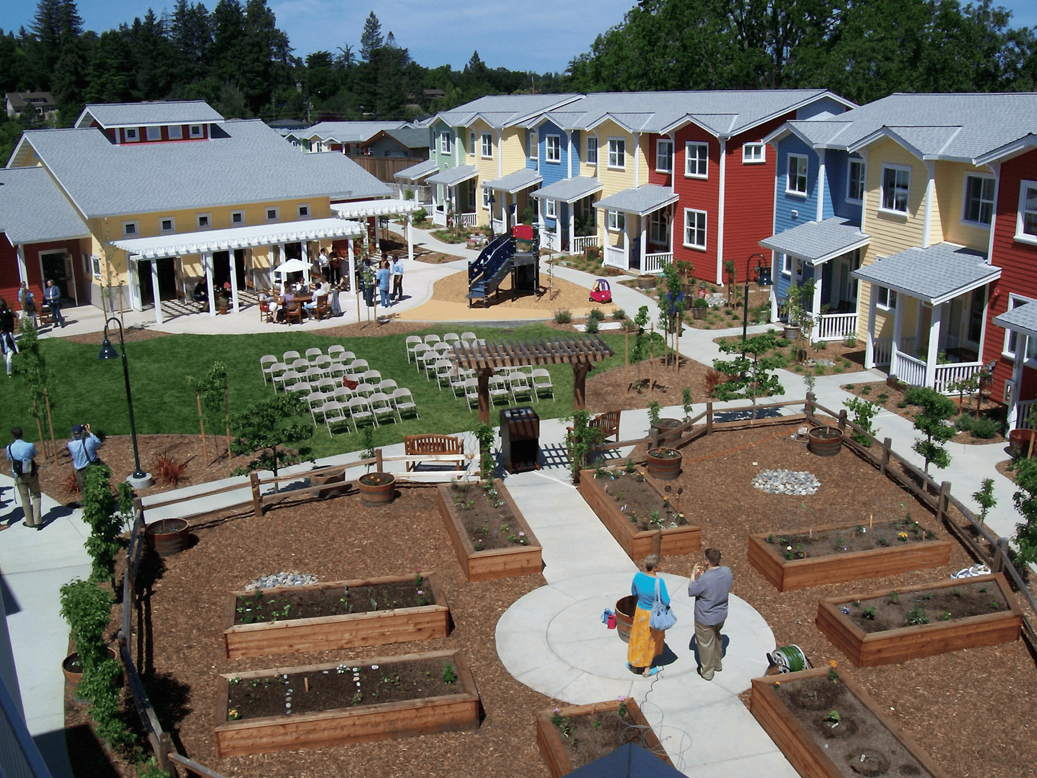 A shared courtyard featuring raised garden beds and colorful row-houses—people walking and tending the space.
