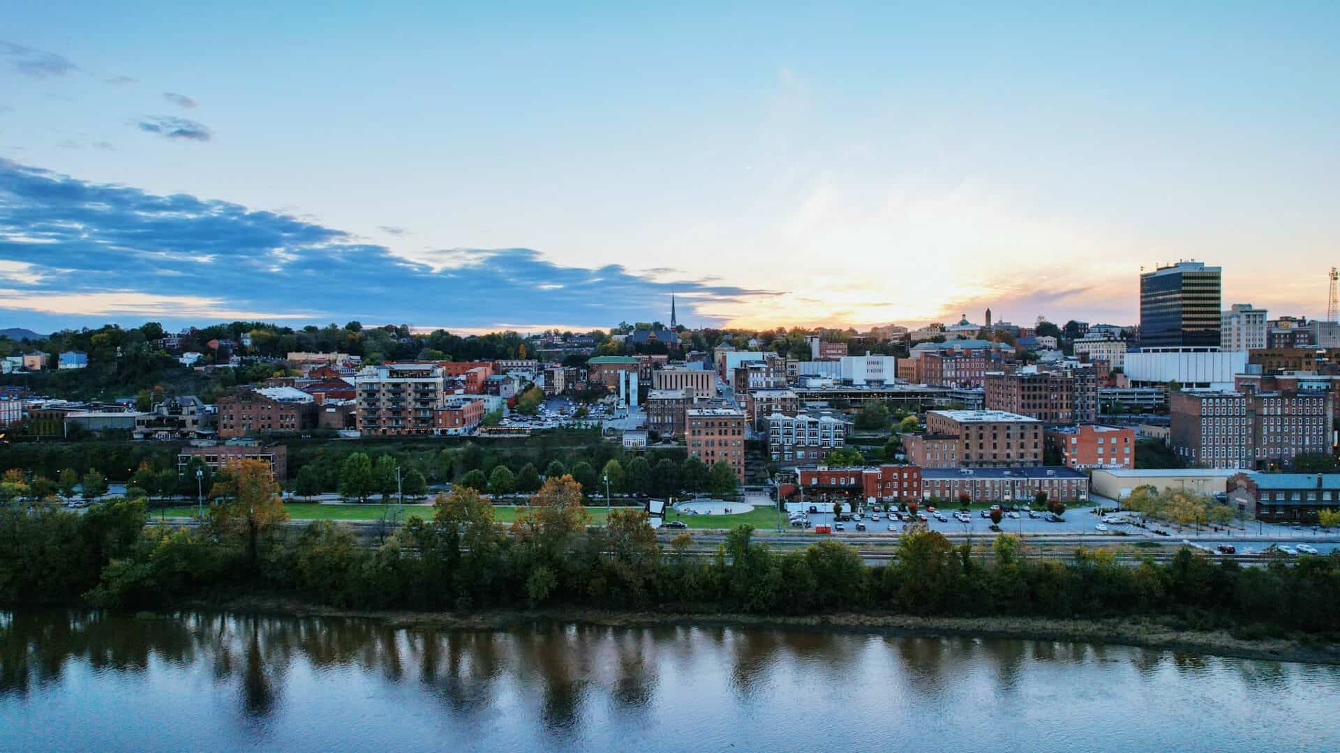 A tranquil view of the James River flowing alongside Lynchburg, with green tree-lined banks and city buildings in the background.