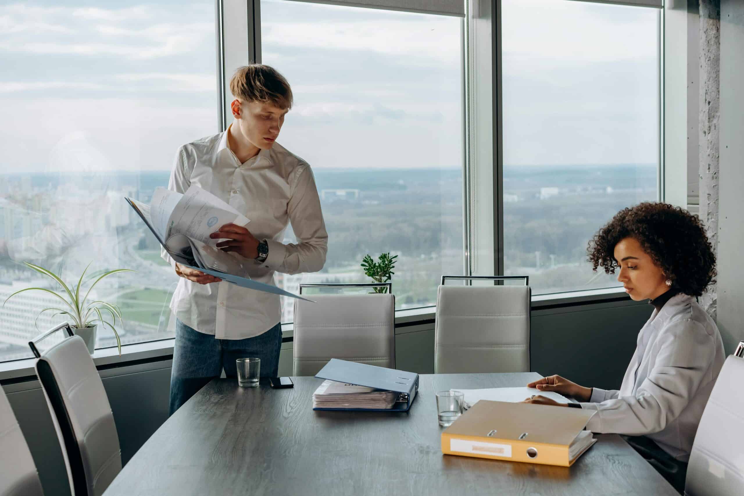 Two people in a modern office reviewing documents at a conference table near large windows.