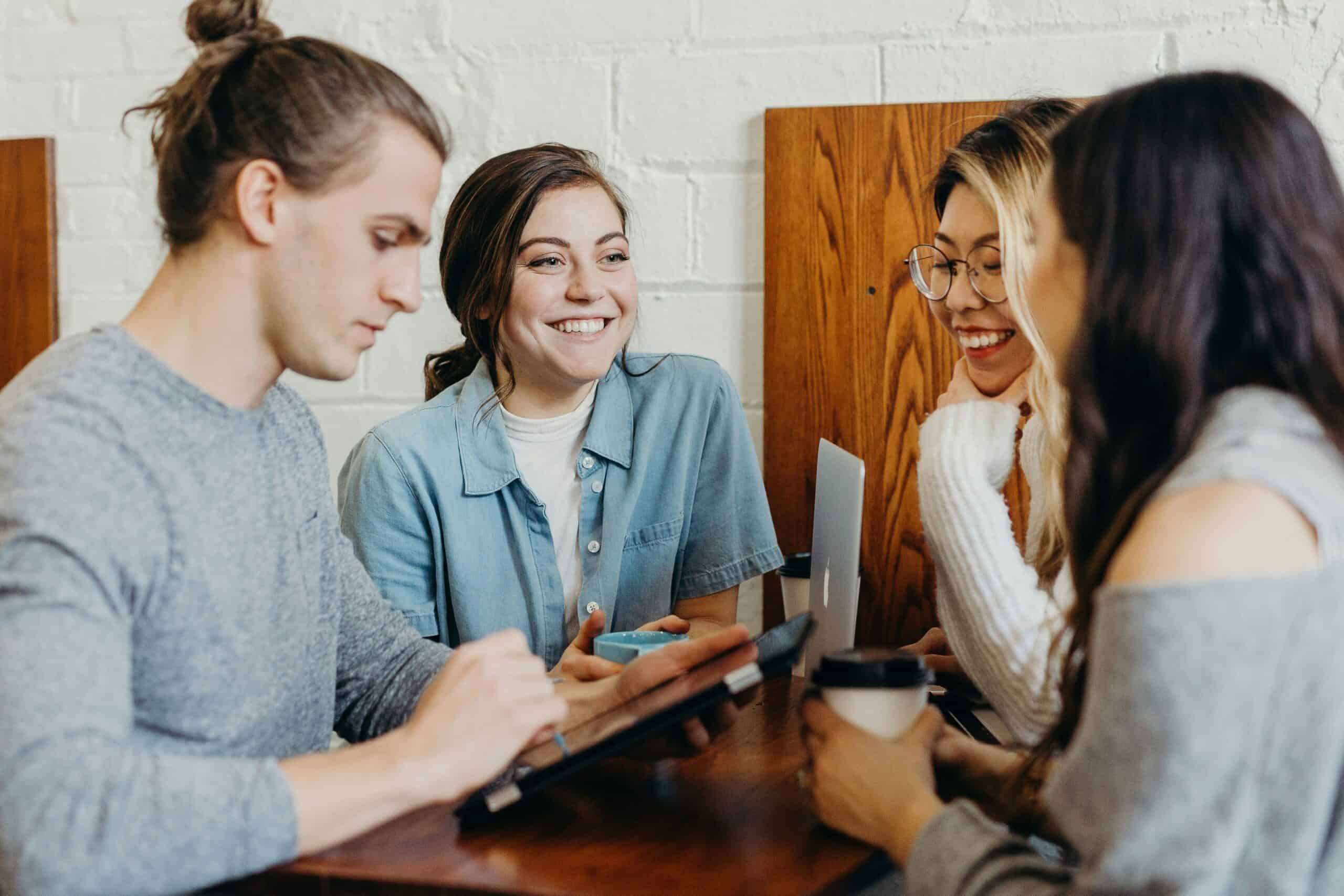 Group of four young people smiling and talking around a table with laptops and coffee.