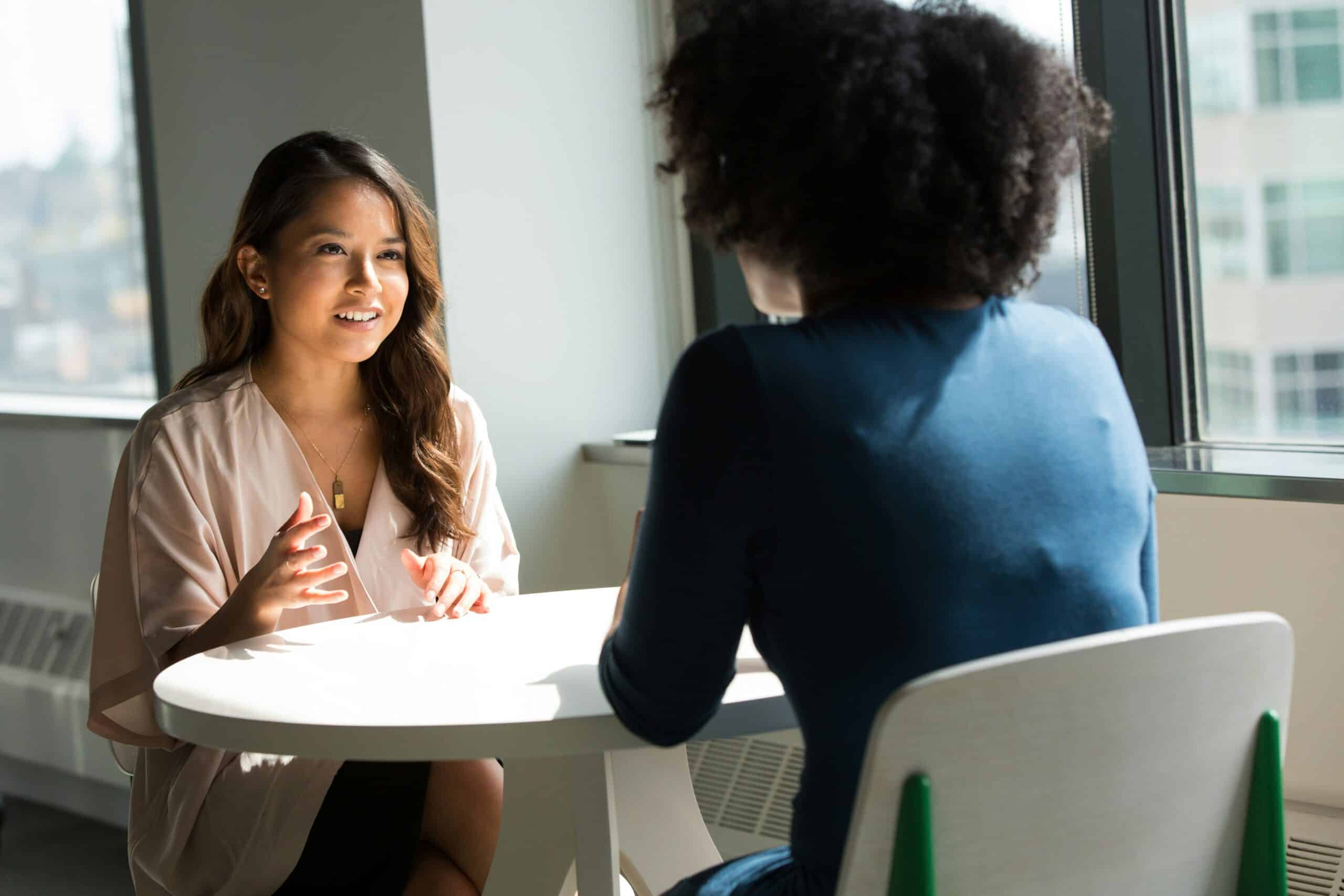 Two women sitting at a table having a conversation in a bright office.