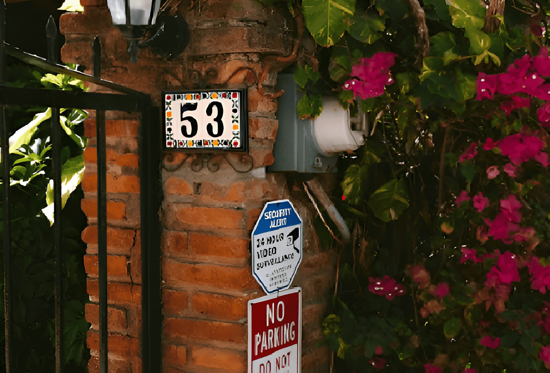 Brick wall with house number 53, security sign, and no parking sign beside pink flowers.