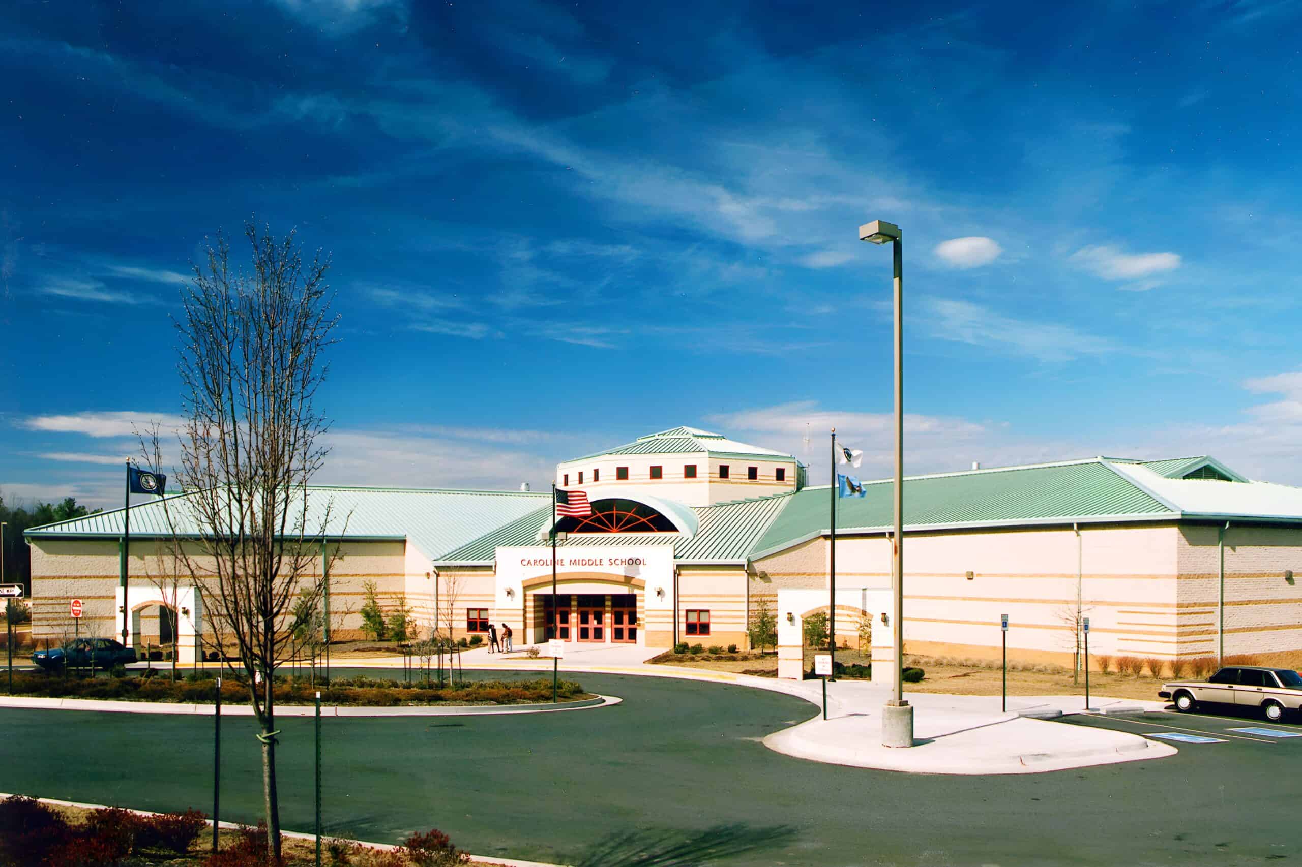 Exterior of Caroline Middle School with a green roof, main entrance, and parking area under a blue sky.